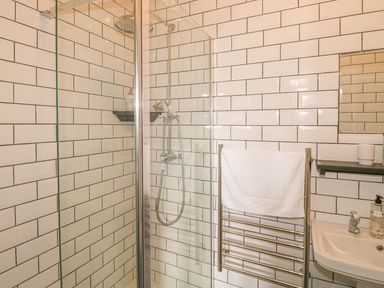 A bathroom with a glass shower enclosure white tiled walls a heated towel rack with a white towel and a sink with a mirror at Nightingale Cottage in Lostwithiel