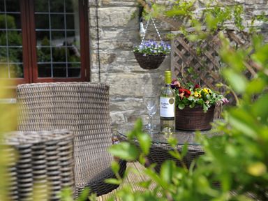 An outdoor patio area with a glass top wicker table and chairs a bottle of white wine two wine glasses and a flowerpot with colorful flowers at Nightingale Cottage in Lostwithiel