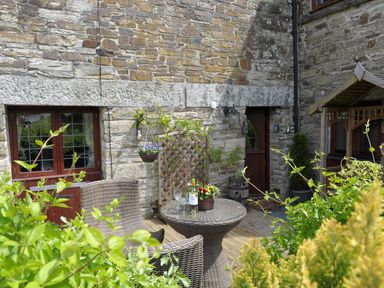 A patio area with a round wicker table and two wicker chairs next to a stone building with a wooden door and window surrounded by plants and flowers at Nightingale Cottage in Lostwithiel