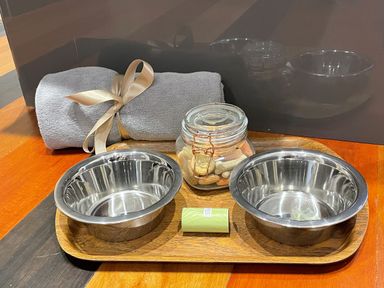 A wooden tray with two empty metal bowls a glass jar with dog biscuits a rolled gray towel with a ribbon and a small green packet at Pams House in Weymouth
