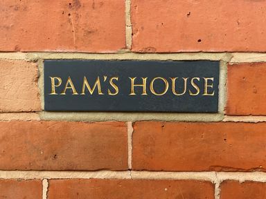 A black plaque with gold text reading PAM'S HOUSE on a red brick wall at Pams House in Weymouth