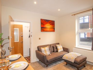 A small living room with a brown sofa and ottoman by a window and a wooden dining table with plates at Pams House in Weymouth