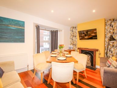 A dining room with a wooden table set for six people a sofa a wall-mounted TV and a window with curtains at Pams House in Weymouth