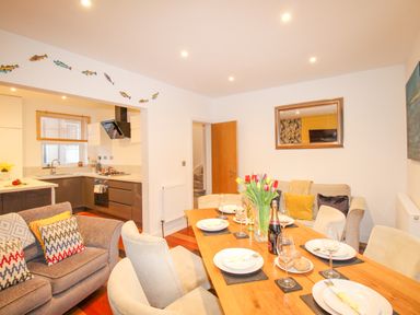A dining area with a wooden table set with plates and glasses adjacent to a kitchen with gray cabinets and a sitting area with sofas at Pams House in Weymouth