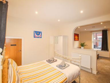 A bedroom with a bed with striped yellow and gray bedding a white wardrobe and a view into a living area with a window and curtains at Pams House in Weymouth