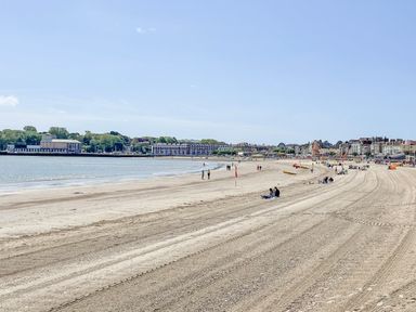 A sandy beach with people sitting and walking near the water and buildings in the background at Pams House in Weymouth