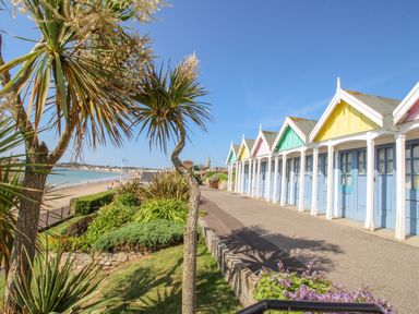 A row of colorful beach huts along a paved pathway next to landscaped greenery and a beach at Pam's House in Weymouth