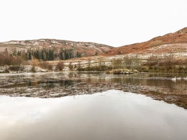 A partially frozen lake with three swans on the water and hills with sparse trees in the background at Ptarmigan Lodge in Glengoulandie near Aberfeldy