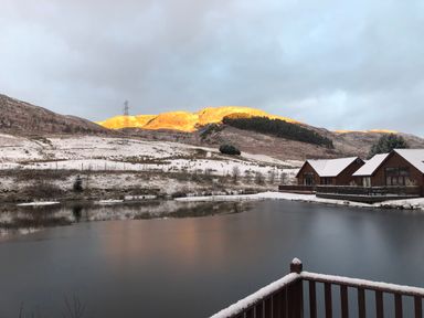 A snowy hillside with wooden cabins next to a frozen lake and a mountain in the background at Ptarmigan Lodge in Glengoulandie near Aberfeldy