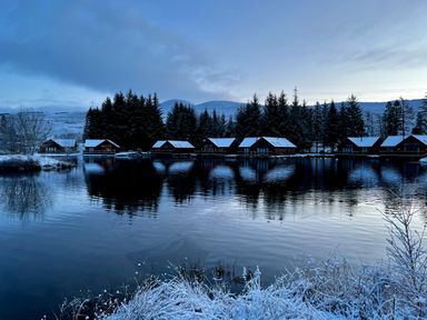 A row of wooden lodges with snow on the roofs beside a lake with trees and snow-covered hills in the background at Ptarmigan Lodge in Glengoulandie near Aberfeldy