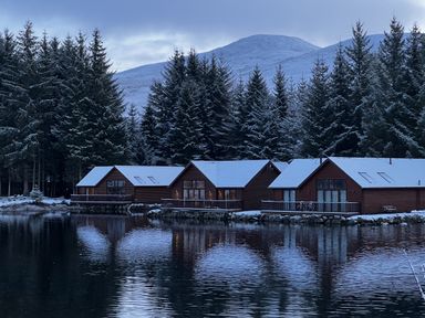 Three cabins with snow on roofs by a lake surrounded by pine trees and mountains at Ptarmigan Lodge in Glengoulandie near Aberfeldy