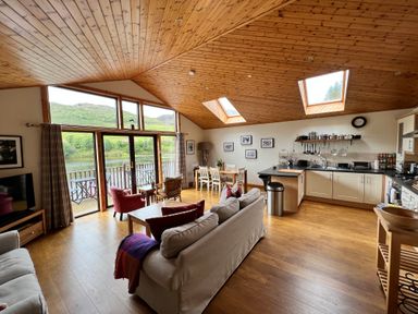 An open living room and kitchen with wooden ceiling and floor large windows and dining table at Ptarmigan Lodge in Glengoulandie near Aberfeldy