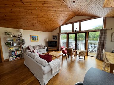 A living room with sofas a bookshelf large windows and a view of a river at Ptarmigan Lodge in Glengoulandie near Aberfeldy