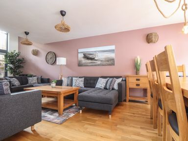 A living room with dark grey sofas wooden coffee table wooden dining chairs and a painting of a boat on a pink wall at Tolcarne in Porthtowan