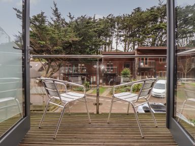 A balcony with two metal chairs overlooking a parking area and wooden buildings with trees in the background at Tolcarne in Porthtowan