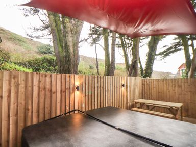 An outdoor area with a hot tub under a red canopy a wooden fence trees and a wooden picnic table at Tolcarne in Porthtowan