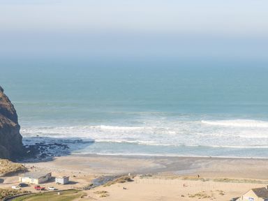 A beach with waves and a rocky cliff near buildings and parked cars at Tolcarne in Porthtowan