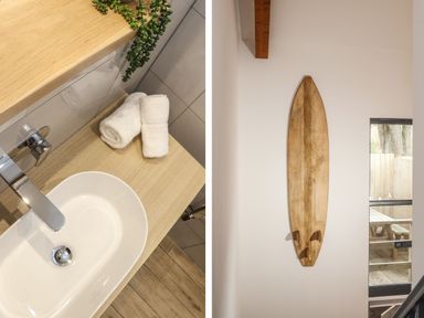 A bathroom sink with rolled towels and a plant next to a wall with a mounted wooden surfboard and a window showing a picnic table outside at Tolcarne in Porthtowan