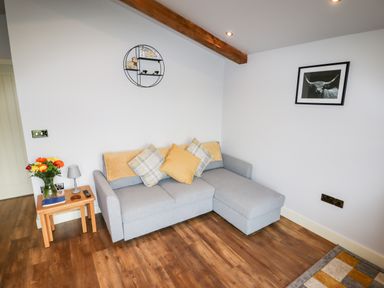 A living room with a grey sectional sofa with cushions a wooden side table with flowers and lamp and wall art at Celyn in Cyffylliog
