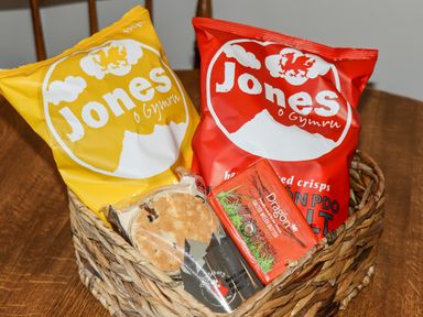 A woven basket containing two bags of Jones crisps and a packet of Dragon salted Welsh butter with oatcakes on a wooden surface at Celyn in Cyffylliog