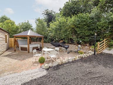 An outdoor patio area with a hot tub under a wooden gazebo a table with four chairs a charcoal grill and surrounding greenery at Celyn in Cyffylliog