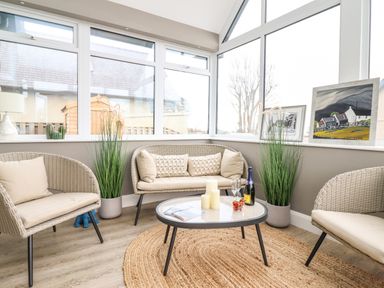 A sunroom with wicker chairs and a wicker loveseat around a round table with candles and a bottle at Bryn Hafod in Dyffryn Ardudwy