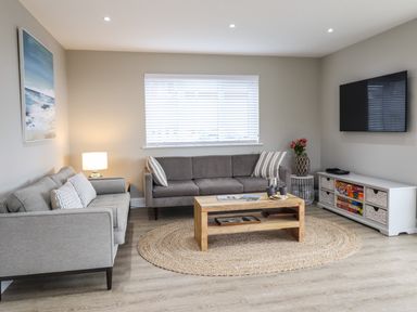 A living room with two sofas a wooden coffee table a round woven rug a wall-mounted television and a cabinet with board games at Bryn Hafod in Dyffryn Ardudwy