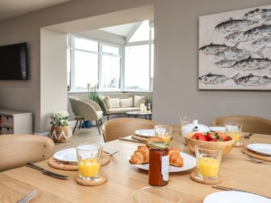 A dining table set with croissants glass of orange juice and a bowl of fruit in a room with seating area and fish painting at Bryn Hafod in Dyffryn Ardudwy