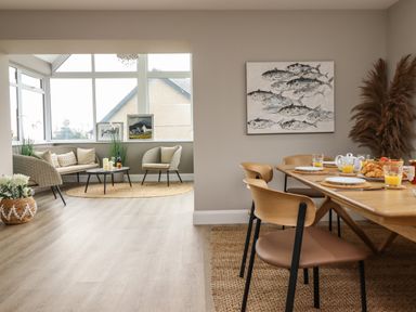 A dining area with a wooden table set for breakfast and a sitting area with wicker chairs and cushions at Bryn Hafod in Dyffryn Ardudwy