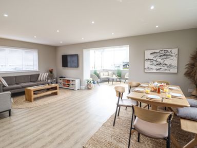 An open living and dining area with a gray sofa, wooden coffee table, wall mounted TV, dining table set with plates and glasses, and a seating nook at Bryn Hafod in Dyffryn Ardudwy