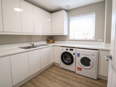 A utility room with white cabinets a stainless steel sink washer dryer under a window at Bryn Hafod in Dyffryn Ardudwy