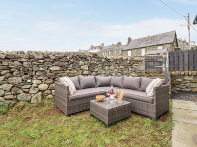 An outdoor seating area with a grey cushioned corner sofa and a small table with drinks on grass by a stone wall at Bryn Hafod in Dyffryn Ardudwy