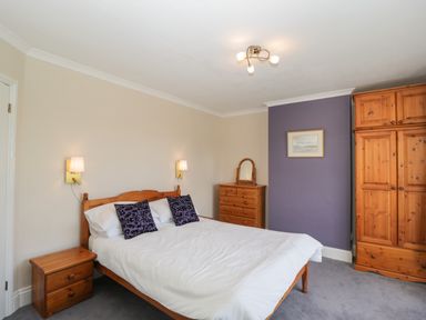 A bedroom with a wooden bed white bedding purple cushions wooden wardrobes a chest of drawers and a bedside table at Caradoc Tutshill near Chepstow