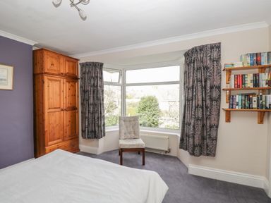 A bedroom with a wooden wardrobe a chair in front of a bay window and a wall-mounted bookshelf at Caradoc Tutshill near Chepstow