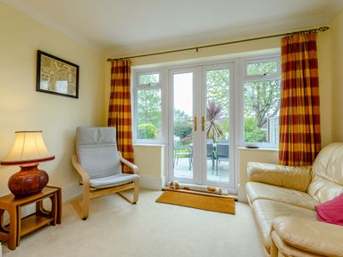A sitting room with a beige sofa a wooden chair with cushion a wooden side table with lamp and door leading to garden at Caradoc in Tutshill near Chepstow