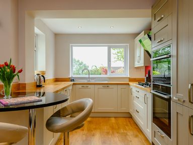 A kitchen with white cabinets wooden countertops a window a sink an oven a gas stove a kettle tulips in a glass vase and two beige stools at Caradoc in Tutshill near Chepstow