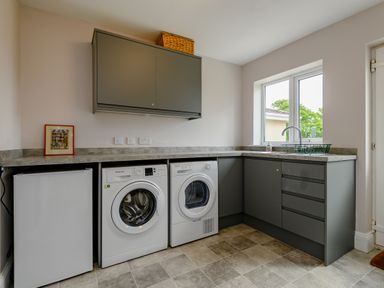 A laundry room with a washing machine and dryer under a countertop with cabinets and a window at Caradoc in Tutshill near Chepstow