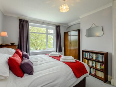 A bedroom with a double bed with red and purple pillows a wooden wardrobe a bookshelf and a window with curtains at Caradoc in Tutshill near Chepstow