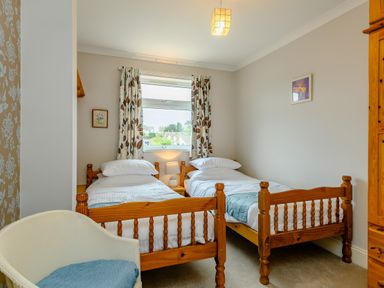 A bedroom with two single wooden beds a window with patterned curtains and a white chair at Caradoc Tutshill near Chepstow
