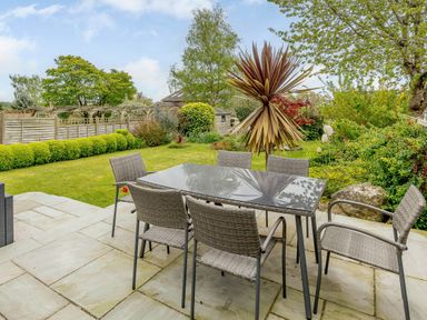 An outdoor patio with a glass table and six wicker chairs overlooking a garden with various plants and a wooden fence at Caradoc Tutshill near Chepstow