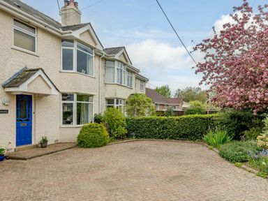 A brick driveway in front of a house with a blue door shrubs and a tree with pink flowers at Caradoc Tutshill near Chepstow