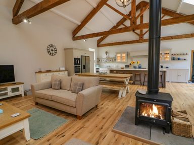 An open plan living area with a wood burning stove a beige sofa a wooden dining table and kitchen with exposed wooden beams at Hollowgill Barn in Sedbergh