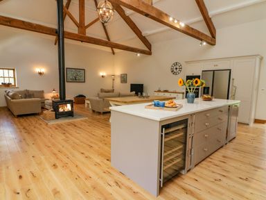 An open-plan living room and kitchen with wooden beams a wood stove sofas and a kitchen island with flowers at Hollowgill Barn in Sedbergh
