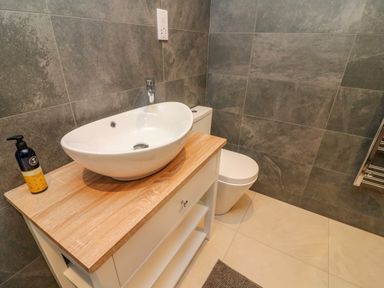 A bathroom with a white sink on a wooden countertop a toilet and grey tiled walls at Hollowgill Barn in Sedbergh