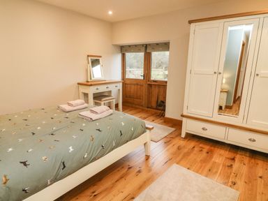 A bedroom with a bed, dressing table with stool, wardrobe with mirror, wooden floor and double doors at Hollowgill Barn in Sedbergh