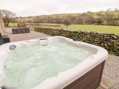 An outdoor hot tub on a gravel patio with stone walls and grassy hills beyond at Hollowgill Barn in Sedbergh
