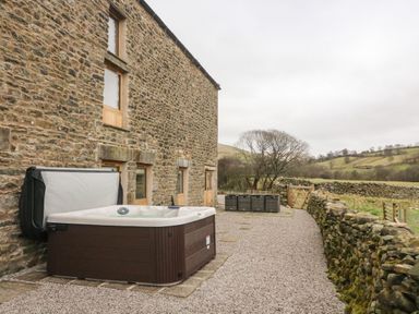 An outdoor area with a hot tub beside a stone building and a gravel path bordered by stone walls at Hollowgill Barn in Sedbergh