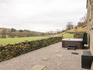 An outdoor area with a hot tub on a gravel surface next to a stone wall and a rural field at Hollowgill Barn in Sedbergh