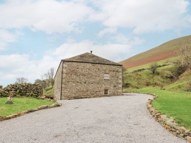 A stone barn building at the end of a gravel driveway with surrounding green hills and stone walls at Hollowgill Barn in Sedbergh