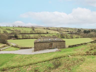 A stone barn with a pitched roof in a rural field with surrounding grass and trees at Hollowgill Barn in Sedbergh